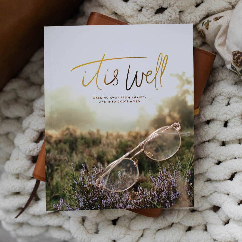 Book titled 'it is well' with a pair of glasses on a nature-themed cover, placed on a textured surface.
