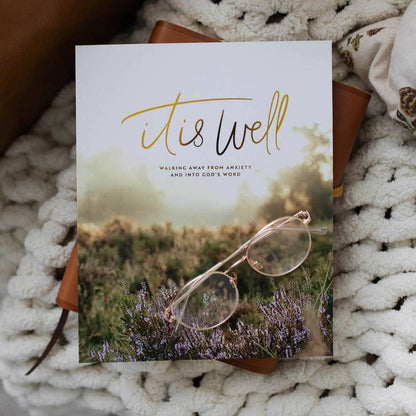 Book titled 'it is well' with a pair of glasses on a nature-themed cover, placed on a textured surface.