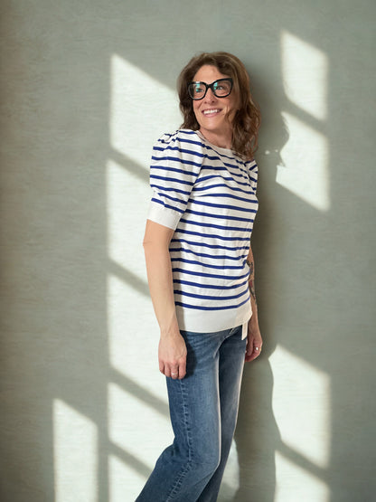 Woman wearing a striped shirt and jeans standing against a wall with sunlight casting shadows.