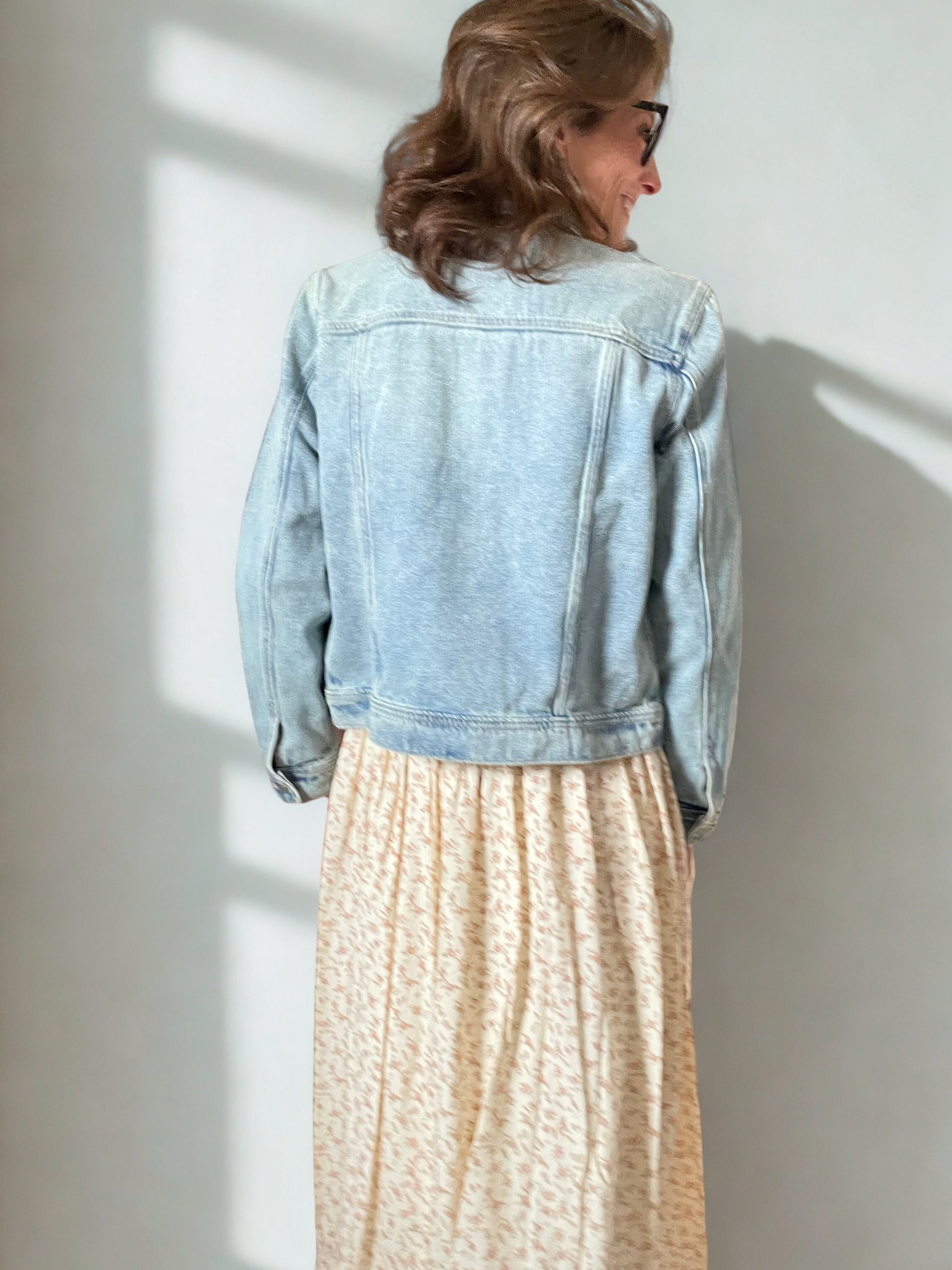 Person wearing a light blue denim jacket and beige skirt standing against a white wall with sunlight casting shadows.
