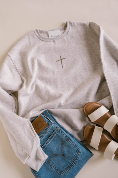 Gray sweatshirt with a cross design, blue jeans, and brown sandals on a light background