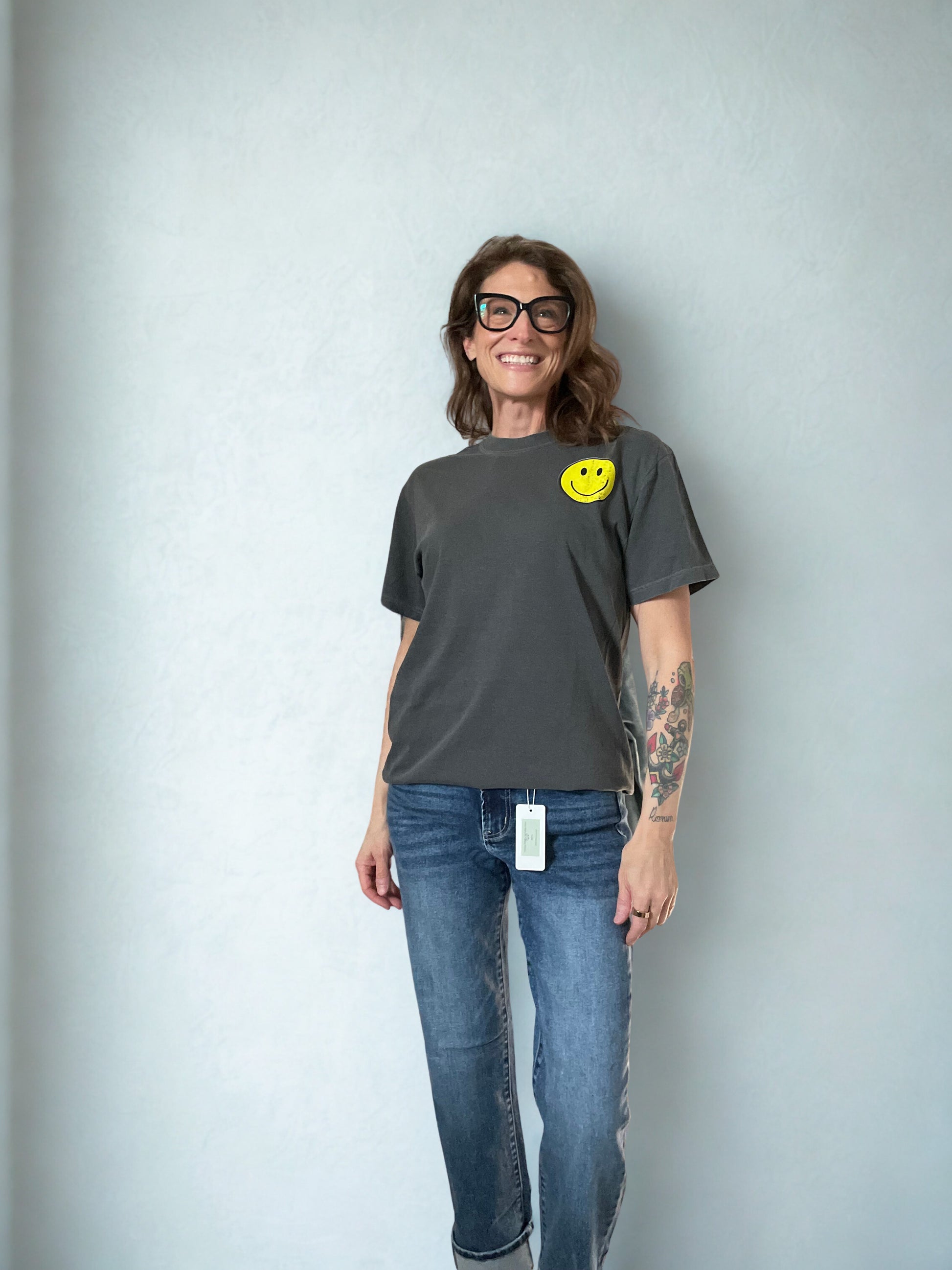 Person wearing a gray t-shirt with a smiley face design and blue jeans against a plain background