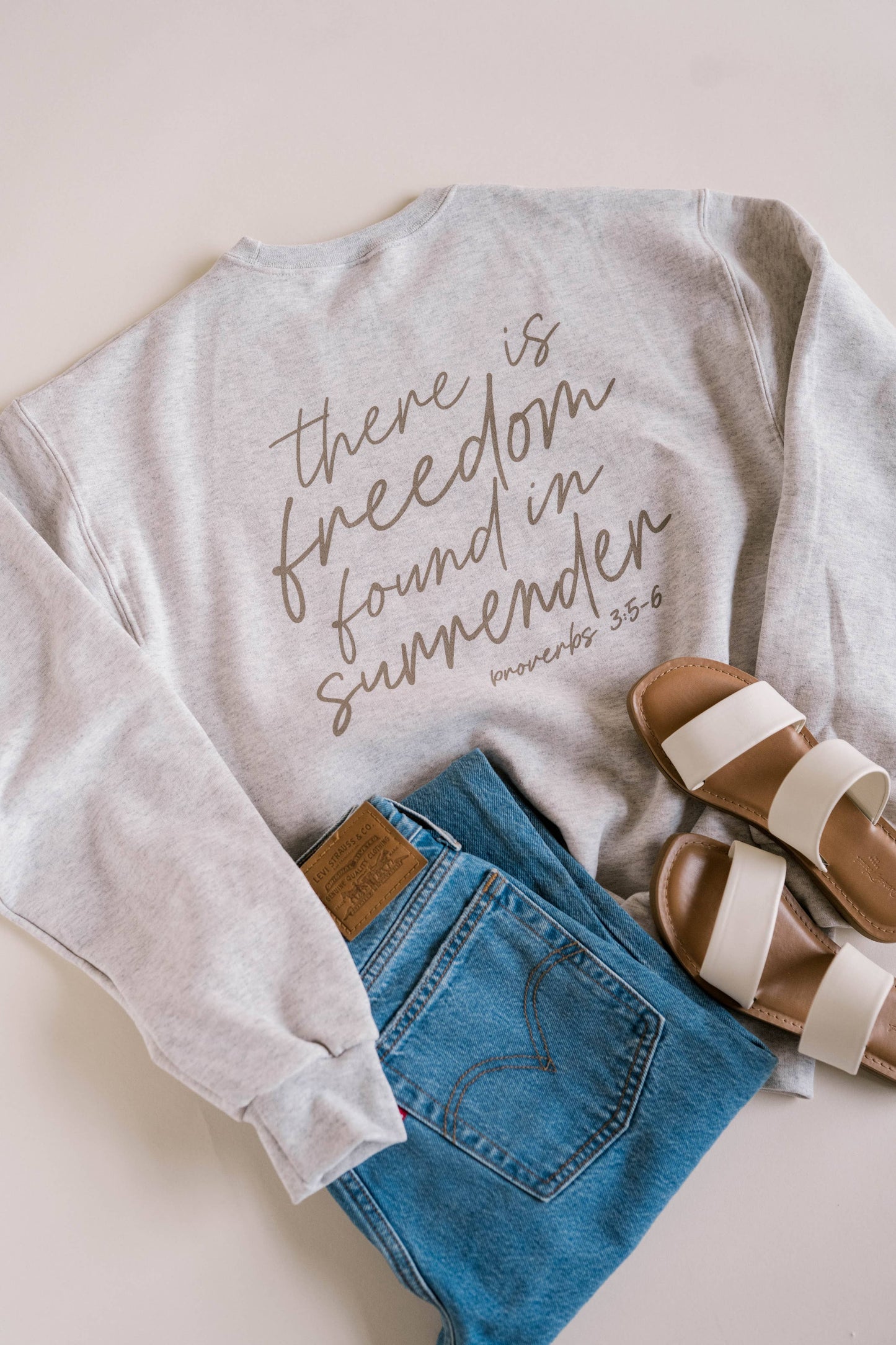 Light gray sweatshirt with text, blue jeans, and brown sandals on a light background