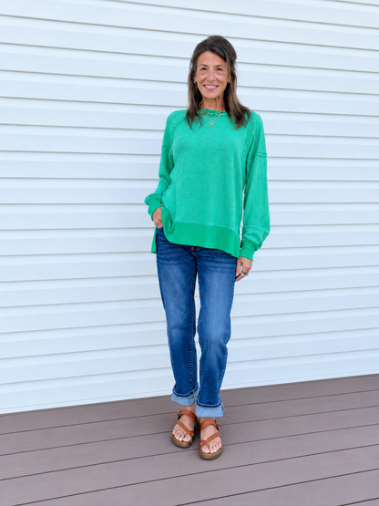 A woman stands on a wooden deck in front of a white siding wall, wearing a bright green long-sleeve knit top, blue jeans, and brown sandals.