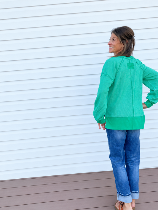 A woman wearing a bright green long-sleeve top and blue jeans stands on a brown deck in front of a white house siding, looking over her shoulder.